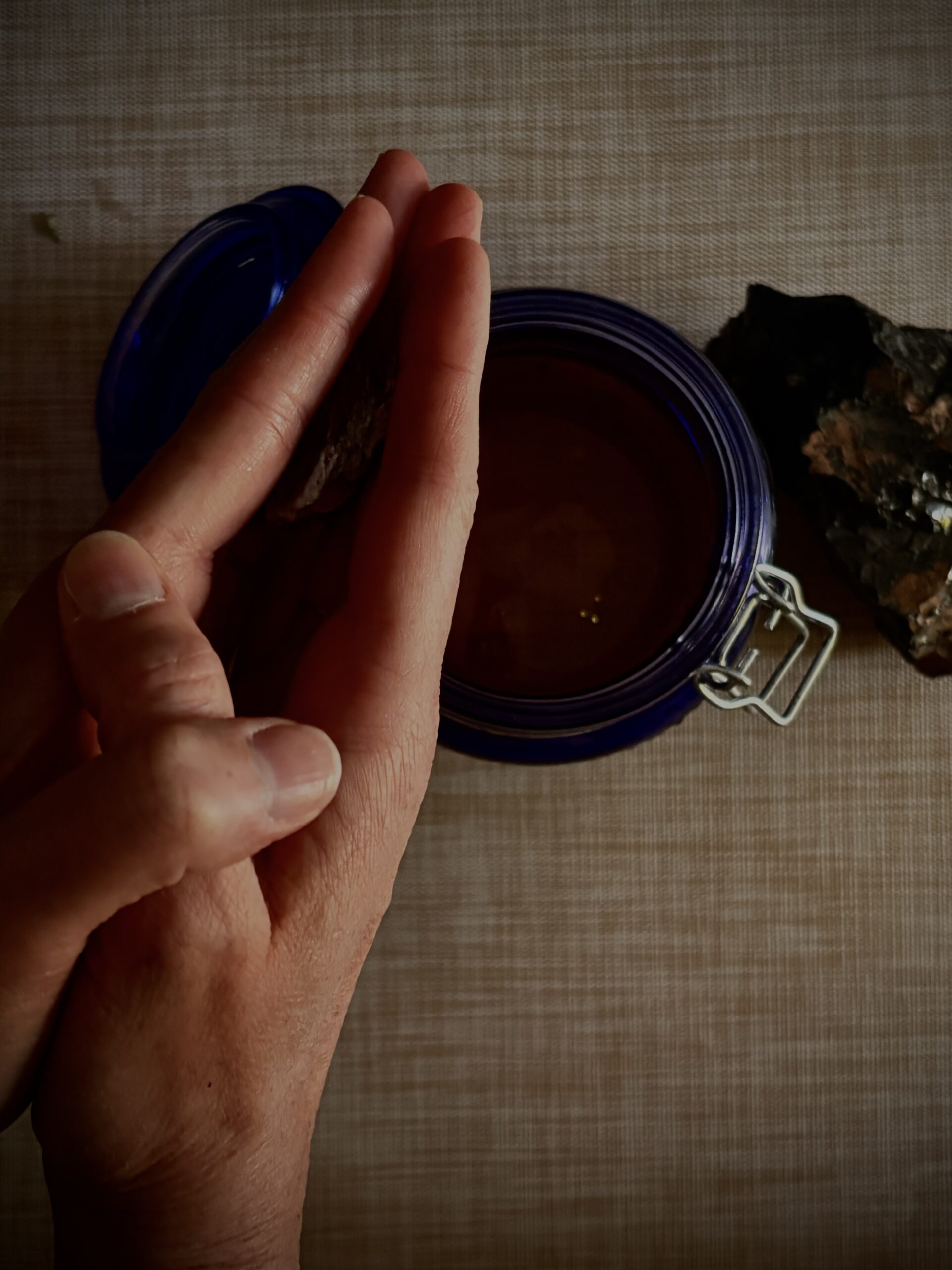 A hand resting beside an open cobalt jar of oil, with a piece of raw stone on the linen alongside it.