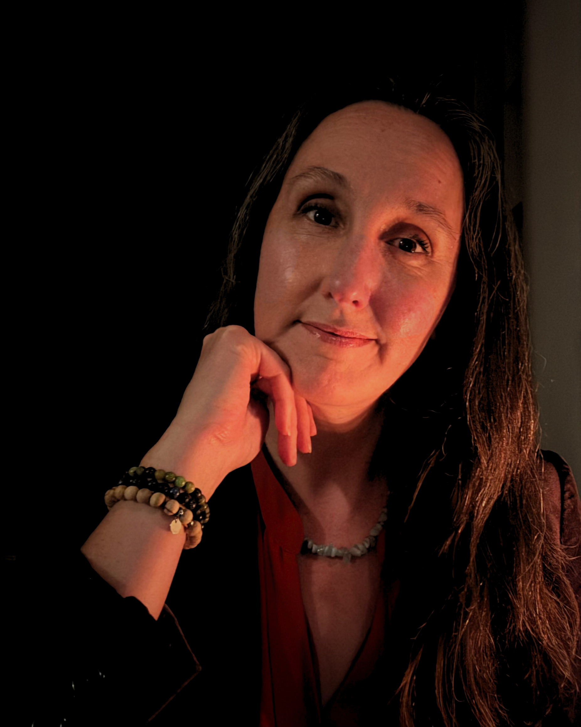 Kristine Cassady in low light, chin resting on her hand, one side of her face lit warm and the rest falling into shadow.